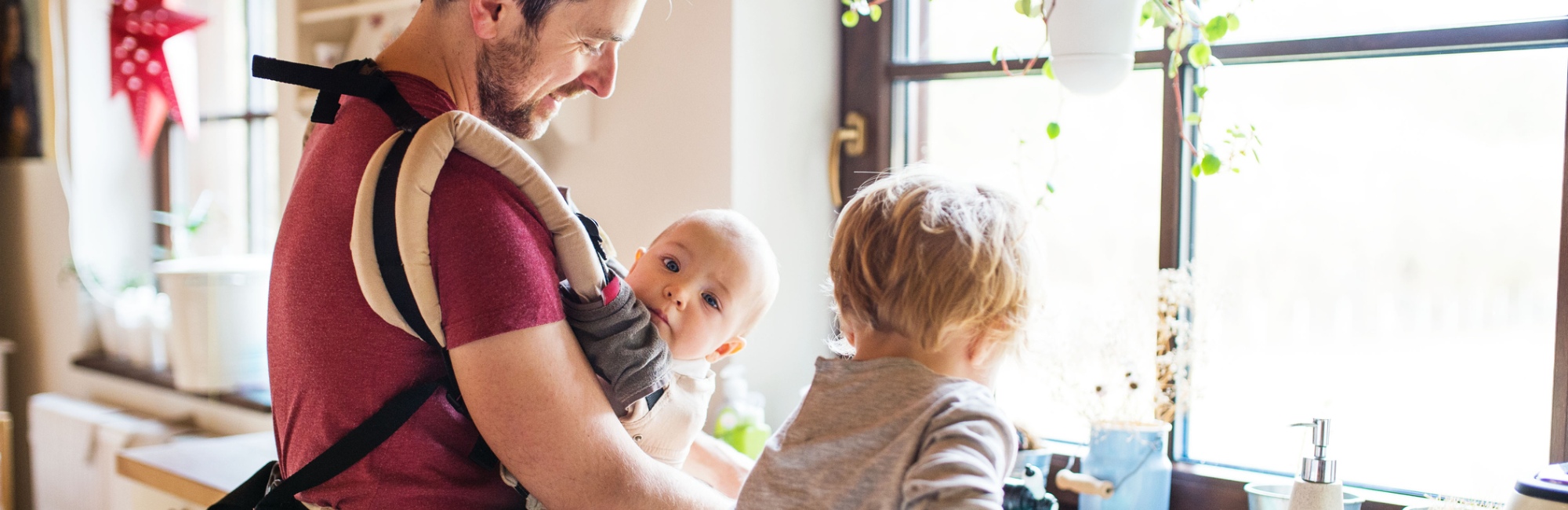 Father carrying baby in daylight in the kitchen while toddler looks on