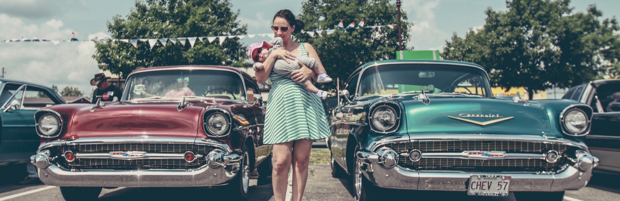 A mother holds her baby in her arms in front of four vintage cars