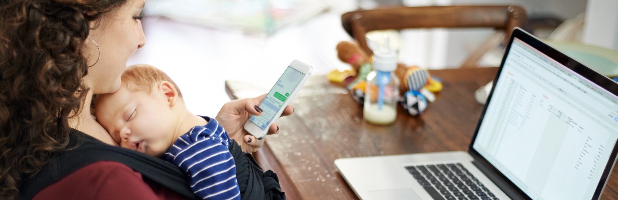 Baby sleeps in sling against mother while she works on her laptop